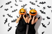 Two Women In Black Dresses Hold Halloween Pumpkins Instead Their Faces On A White Background With Bats And Spiders