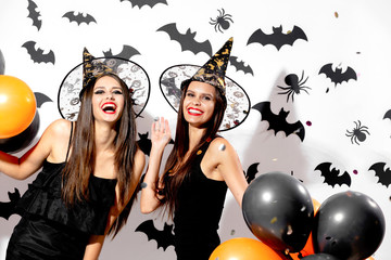 Two charming young women in witches hats hold black and orange balloons on a white background with black bats. Confetti around