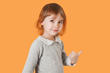 Portrait of smiling little baby girl indicates with thumb to an side isolated on a orange background.