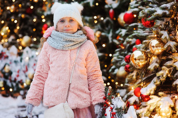 Wall Mural - happy child girl playing outdoor on the walk in snowy winter city decorated for new year holidays. Trees with christmas lights on background