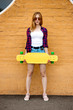© Leika production - A girl wearing sunglasses, checkered shirt and denim shorts is standing in front of the brick wall and holding a yellow longboard in front of her. Cool style.