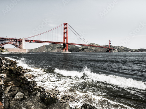 Fotografie, Obraz Golden Gate bridge with waves crashing against rocks in the foreground along the San Francisco Bay in California