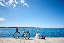 Back View Of Young Woman In White Closing, Sunhat And Sunglasses Sitting At Bicycle On Stony Sidewalk Under Clear Blue Sky On Sparkling Clear Water Background. Tourism And Vacations Concept.