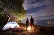 © anatoliy_gleb - Back view of young tourist couple, athletic man and slim woman standing holding hands near tourist tent by fire on evening sky and sea water background. Active lifestyle and loving relations concept.