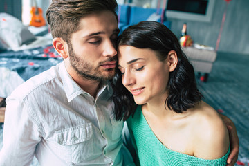 Beautiful young tender lovely couple are kissing and hugging each other while sitting on the floor of bedroom with romantic background. St Valentine's day. Anniversary. Date. Love concept