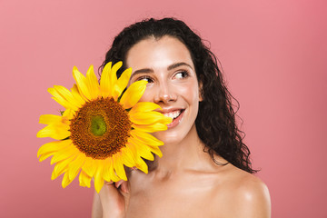 Beauty photo of caucasian nude woman 20s with long hair laughing and holding yellow sunflower, isolated over pink background