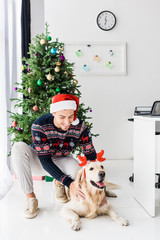 smiling man in santa hat stroking golden retriever dog in deer horns on christmas eve