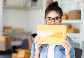 Portrait of a pretty Asian young girl hiding behind box and look at camera with smile with people in background.Teenager business owner work at home. Small business owner concept