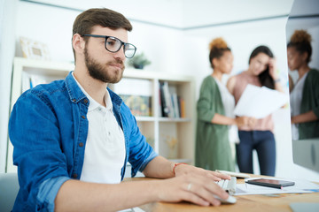 Portrait of young focused businessman sitting at desk, using computer and enjoying work in modern office