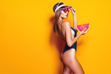 Beautiful Young Female In Cap, Sunglasses And Swimsuit Hold Slice Of Watermelon And Looking Away While Standing On Yellow Background. Copyspase