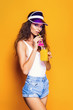 © zamuruev - Sexy Woman in white swimsuit and blue jeans shorts, trendy visor holding glass of fresh drink while standing on yellow, orange hot background