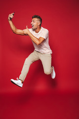 Poster - Full length photo of handsome man in striped t-shirt jumping and taking selfie on smartphone, isolated over red background