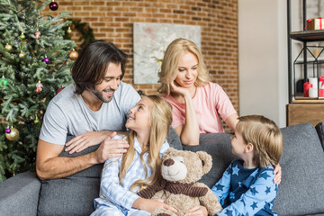 happy parents looking at adorable smiling children in pajamas sitting in sofa at christmas time