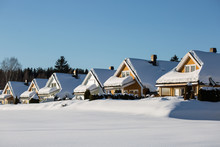 A Row Of Little Wooden Houses Covered With A Thick Snow Layer, Deep Untouched White Snow Around