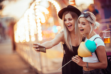 Wall Mural - Happy female friends in amusement park eating cotton candy and taking selfie.Two young women enjoying a day at amusement park. 