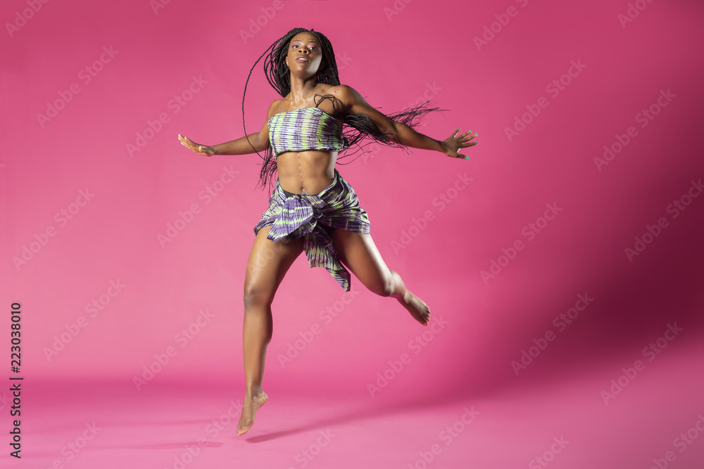 Beautiful African Black girl wearing traditional colorful African outfit does a dramatic dance move against a colorful pink background - obrazy, fototapety, plakaty 