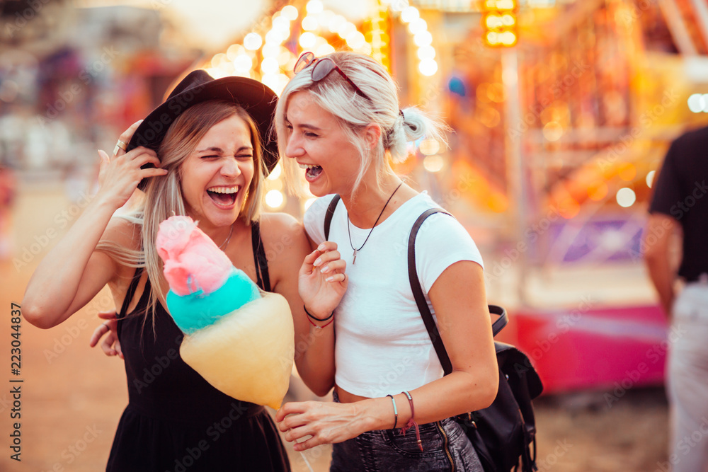 Shot of happy female friends in amusement park eating cotton candy. Two young women enjoying a day at amusement park.  - obrazy, fototapety, plakaty 