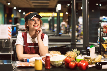 Wall Mural - Young cheerful female cashier in uniform joyfully looking aside with products on cash line near while working in modern supermarket