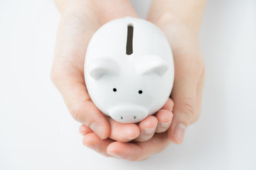 Wall Mural - Savings and invest for future growth. top view of young female hands tender holding white piggy bank on white table in soft tone