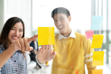 Young successful creative man and woman smile and brainstorm on project at office pointing hand or finger to yellow post note or sticky note. Close up view couple sharing idea on glass wall concept.