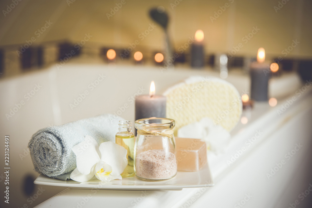 White ceramic tray with home spa supplies in home bathroom for relaxing rituals. Candlelight, salt soap bar, bath salt in jar, massage, bath oil in bottle, blue rolled towel, natural sponge.  - obrazy, fototapety, plakaty 