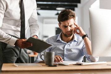 Wall Mural - tired bored displeased man sitting in office working with computer near his colleague who showing cl