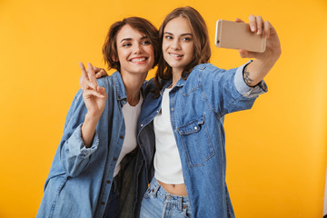 Sticker - Emotional young women friends posing isolated over yellow background take a selfie by mobile phones.