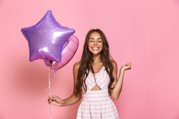 Wall Mural - Portrait of stylish caucasian girl 20s with long hair wearing dress smiling and holding two balloons, isolated over pink background