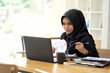 © bongkarn - Attractive young Asian wearing dark hijab working paperwork with her laptop.