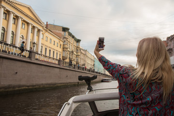 Woman with fluttering hair sails on tour boat on river, making selfie in the city at sunset.