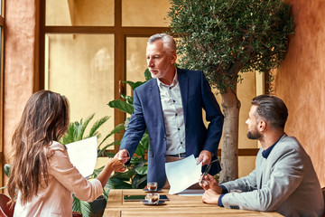 Wall Mural - Business lunch. Three people in the restaurant sitting at table making deal senior man shaking hands with young woman. Agreement concept