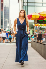 Young Eastern European Woman with long brawn hair traveling in New York City, wearing blue sleeveless, v neck, jumpsuit, black leather shoes, walking on busy street in middletown of Manhattan..