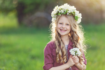smiling little girl with long blond hair and wreath of flowers in a blooming apple orchard