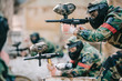 selective focus of paintball player in protective mask aiming with marker gun and his teammate behind outdoors