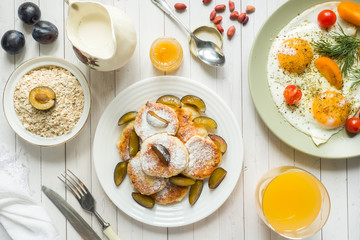 Wall Mural - Concept of Breakfast. Fried eggs, cottage cheese pancakes, plums and oatmeal with milk, orange juice on the table