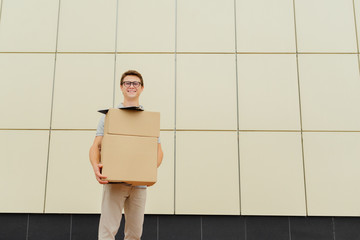 Smiling attractive delivery guy looking at the camera and holding two boxes of parcels, standing against the modern wall, outdoors.