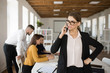 © Anton - Young serious business woman in eyeglasses and shirt thoughtfully talking on cellphone in office with colleagues on fuzzy background