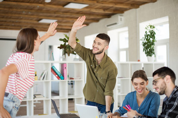 Young joyful man in shirt happily giving  high five to colleague while spending time at work. Group of creative guys working together in modern office