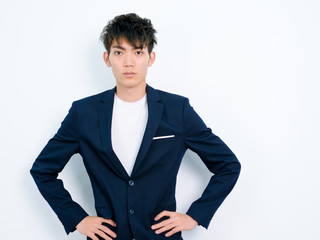 Portrait of handsome Chinese young man in dark blue leisure suit posing against white wall background. Two hands on waist with confident expression.