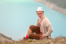 Beautiful Portrait Of A Handsome Young Man In Traditional Clothing With Lederhosen And Checkered Shirt In Summer In The Alps Near Grimentz, Switzerland, With Blue Lake Lac De Moiry In The Background