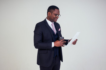 Handsome young African man in formal black suit holds a plastic paper cup with hot coffee cocoa (tea) to go and paper documents in hand . businessman on coffee break after meeting on white background 