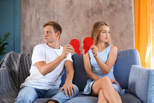 Young Couple Holding Halves Of Broken Heart On Sofa At Home. Relationship Problems