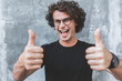 © iuricazac - Horizontal shot of handsome happy smiling male with curly hair, wears sepctacles posing for social advertisement with thumbs up on gray concrete wall with copy space for your text.
