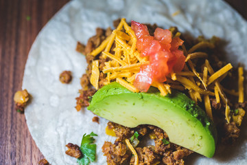 A close up of a vegan taco with ground meat, avocado, cheese, and diced tomato on a wood board.