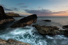 A Long Exposure Of Blurred Waves Swirling And Crashing Over Rugged Rocks On The Rocky Cornish Coastline In Cornwall, UK.