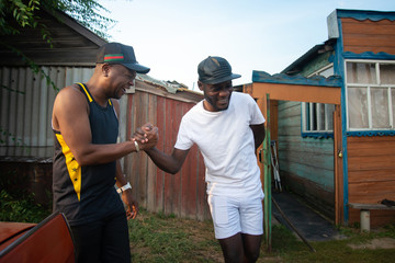 Two black men laughing and shaking hands in the background of village house