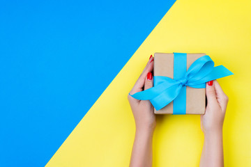 woman hands holding gift wrapped and decorated with blue bow on blue and yellow background with copy space