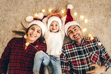 Christmas. Family. Happiness. Top view of dad, mom and daughter in Santa hats looking at camera and smiling while lying on the floor at home