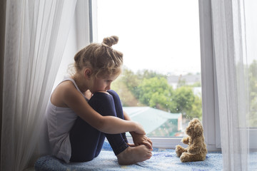 sad litttle girl sits on a window sill and looks at a bear