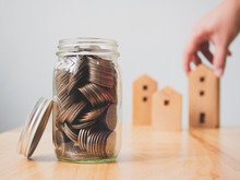 Property Investment Real Estate And House Mortgage Financial Concept. Money Coins In Jar With Hand Holding Wooden Home On Table